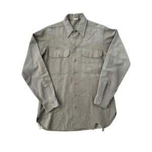 May include: A long-sleeved, button-down shirt in a muted olive green color. The shirt features two chest pockets, a classic collar, and buttoned cuffs. The fabric appears to be a durable, woven material. The bottom hem has some wear and tear.