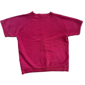 Pode incluir: Uma camisola magenta de manga curta com gola redonda. As mangas raglan estendem-se até meio do braço superior. A bainha inferior e a gola são caneladas. O tecido parece ser um material de malha macia.