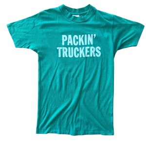 Puede incluir: Una camiseta de manga corta y cuello redondo en un vibrante color verde azulado. La parte delantera de la prenda presenta el texto "PACKIN' TRUCKERS" impreso en una fuente sans-serif clara y llamativa, centrada en la zona del pecho.
