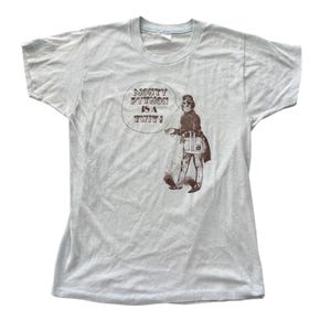 May include: A light gray short-sleeve t-shirt featuring a dark brown graphic of a person in Victorian-style clothing holding a bag. A speech bubble above the figure contains the text, "MONTY PYTHON IS A TWIT!" in a bold, dark brown font.
