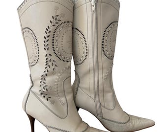 May include: A pair of off-white knee-high boots featuring a pointed toe, a slim stiletto heel, and a full-length side zip. The boots are decorated with intricate laser-cut floral and circular patterns along the shaft and toe area.