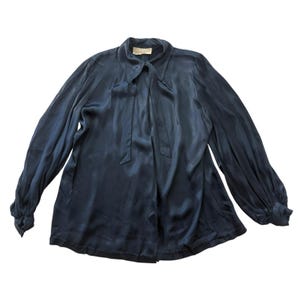 May include: A dark navy blue long-sleeve blouse with a satin-like finish, featuring a pointed collar, a front neck tie detail, and gathered cuffs. The garment has a loose, flowing silhouette with visible fabric folds and a label at the neckline.