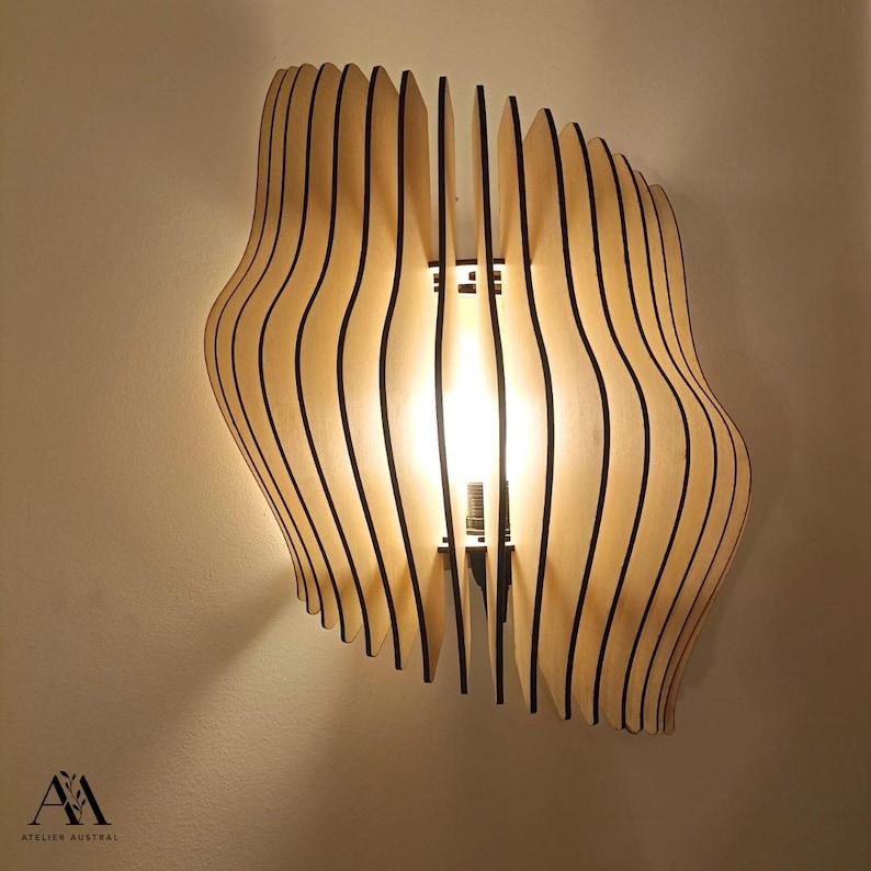 May include: A wooden wall sconce with a unique, layered design. The light fixture features curved, vertical wooden slats with black edges, creating a wave-like pattern. The light shines through the slats, illuminating the room. The text "ATELIER AUSTRAL" is visible.