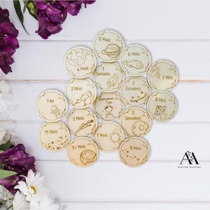 May include: A collection of wooden discs with engraved space-themed illustrations and text. Each disc features a different design, including planets, rockets, and astronauts, along with numbers and time units like "Mois" and "Semaines". The discs are arranged on a white surface.