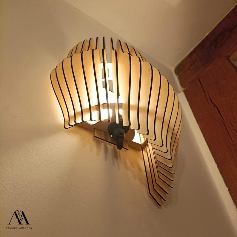 May include: A wooden wall sconce with a layered, curved design. The sconce has a light tan color with black accents and a dark metal light fixture. The light shines through the wooden slats, creating a warm glow. The sconce is mounted on a white wall.