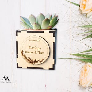 Peut inclure: Un petit pot en bois avec une plante succulente. La boîte a un design décoratif et une pièce de bois circulaire avec le texte "Mariage Laure & Théo" et la date 27/09/2025. Le pot est brun clair avec des accents noirs.