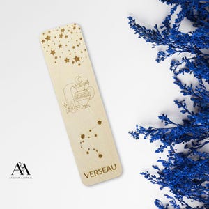 May include: A wooden bookmark with etched celestial designs. The top features stars, the middle shows an Aquarius symbol, and the bottom has the word "VERSEAU". The bookmark is light brown and has a rectangular shape. Blue dried flowers are in the background.