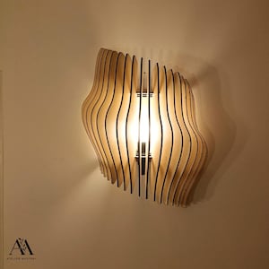 Peut inclure: Applique murale en bois au design ondulé unique. Le luminaire est composé de fines lattes de bois verticales aux bords noirs, créant un effet de superposition. La lumière traverse les lattes, éclairant la pièce. L'applique est fixée au mur.