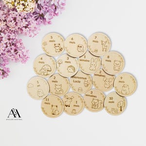 Può includere: Dischi di legno per pietre miliari con illustrazioni incise di animali e numeri. I dischi sono disposti su una superficie bianca, con un bouquet di fiori viola sullo sfondo. I dischi sono per il primo anno del bambino.