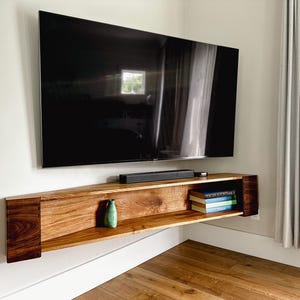 May include: A wall-mounted, wooden entertainment unit with a large, black television above it. The shelf has a natural wood grain finish and contains a small green vase and a stack of books. The TV is approximately 152 cm.