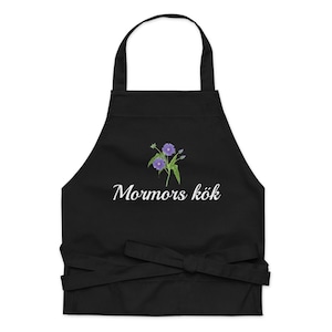 May include: Black apron with a floral design and the text "Mormors kök" in white script. The apron features a purple flower illustration and a waist tie.