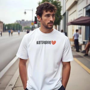 Puede incluir: Camiseta blanca con el texto "KATTPAÑPA" en negro y un corazón rojo. La camiseta es de manga corta y parece estar hecha de un material suave. La persona está de pie en una acera.