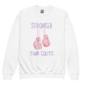 May include: White sweatshirt with the text "STRONGER THAN COLITIS" in purple. The design includes two pink boxing gloves. The sweatshirt is a Gildan brand.