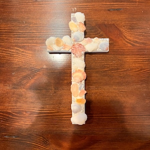 May include: A white wooden cross decorated with various seashells and small pearls. The shells are in shades of white, tan, and pink. The cross is displayed against a dark wood grain background.