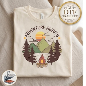 May include: A cream-colored t-shirt featuring a graphic print with the text ADVENTURE AWAITS. The design includes a sunset over green mountains, dark brown pine trees, and a campfire with logs. A gold circular badge reads HIGH QUALITY DTF TRANSFER.