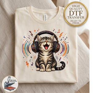 May include: A cream-colored t-shirt featuring a graphic of a tabby kitten wearing large headphones with its mouth open. The design includes colorful sound waves, musical notes, and stars. A gold circular badge reads High Quality DTF Transfer.