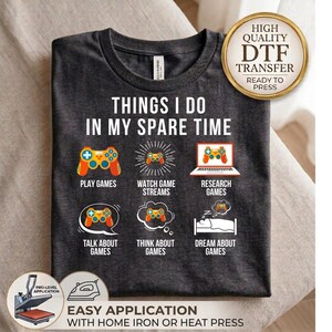 May include: A dark charcoal gray t-shirt featuring the white text THINGS I DO IN MY SPARE TIME above six icons representing gaming activities: a controller, a stream, a laptop, a speech bubble, a thought bubble, and a person sleeping in a bed.
