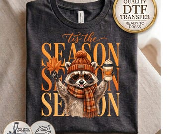 May include: A dark charcoal gray t-shirt featuring a graphic of a raccoon wearing a brown knit hat, heart-shaped sunglasses, and a plaid scarf. The raccoon holds an orange maple leaf and a coffee cup. The text reads Tis the SEASON.