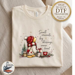 May include: A cream-colored t-shirt featuring a graphic of a bear wearing a red knit hat and scarf, standing next to a pine tree, gifts, and a red mug. The text reads: Small moments, big blessings, warm heart. A gold circular badge is in the corner.