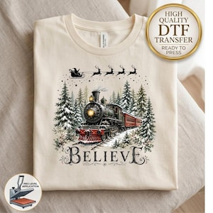 May include: A cream-colored t-shirt featuring a graphic of a black steam locomotive traveling through a snowy forest. Above the train, Santa Claus and his reindeer fly in silhouette. The word BELIEVE is printed in black below the train.