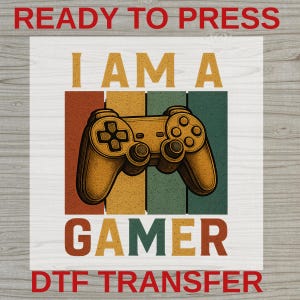 May include: A rectangular DTF transfer sheet featuring the text READY TO PRESS at the top and DTF TRANSFER at the bottom. The center graphic displays a yellow game controller over vertical stripes in red, yellow, and green with the text I AM A GAMER.