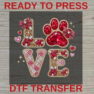 May include: A ready to press DTF transfer featuring the word LOVE with a paw print replacing the letter O. The design is decorated with red and pink gemstones, gold filigree accents, and small bows. The text READY TO PRESS DTF TRANSFER is visible.