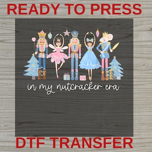 May include: A DTF transfer sheet featuring a festive illustration of two nutcrackers, two ballerinas, and a mouse character, surrounded by Christmas trees and gifts, with the white text "in my nutcracker era" printed below the characters.