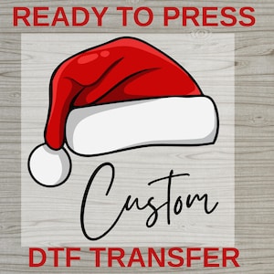 May include: A red Santa hat with a white brim and a white pom-pom. The word "Custom" is written in a cursive font below the hat. The words "READY TO PRESS" and "DTF TRANSFER" are in red block letters.