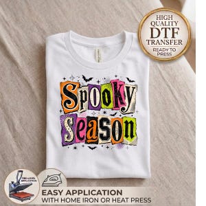 May include: A white t-shirt featuring a colorful graphic print with the text "SPOOKY SEASON" in a distressed, block-letter style. The design includes orange, lime green, and purple accents, spider webs, bats, and stars on a white background.