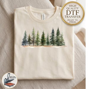 May include: A cream-colored t-shirt featuring a watercolor-style graphic of a forest line with various green pine trees. A gold circular badge in the top right corner reads HIGH QUALITY DTF TRANSFER READY TO PRESS. A small icon in the bottom left shows a heat press.