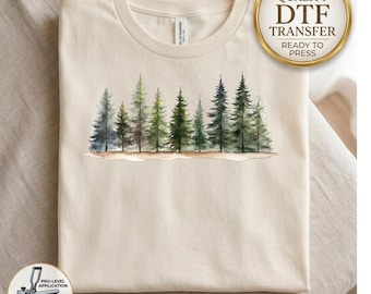 May include: A cream-colored t-shirt featuring a watercolor-style graphic of a forest line with various green pine trees. A gold circular badge in the top right corner reads HIGH QUALITY DTF TRANSFER READY TO PRESS. A small icon in the bottom left shows a heat press.