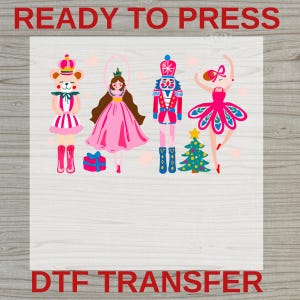 May include: A DTF transfer sheet featuring the text READY TO PRESS and DTF TRANSFER. The graphic displays a bear in a crown, a ballerina in a pink dress, a nutcracker, and a dancer in a pink tutu next to a small Christmas tree and a gift box.
