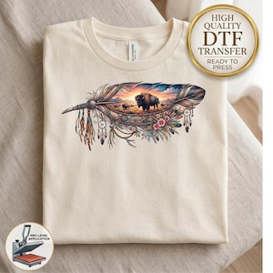 May include: A cream-colored t-shirt featuring a large graphic of a feather with intricate patterns, hanging beads, and feathers. Inside the feather is a landscape scene with a bison standing in a field at sunset, accented by small pink flowers.