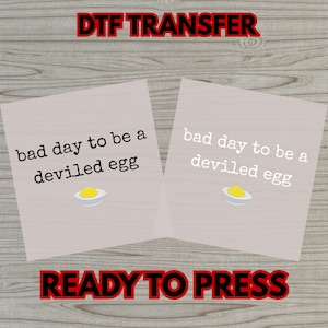 May include: Two light gray DTF transfer sheets with the text "bad day to be a deviled egg" and a deviled egg graphic. The words "DTF TRANSFER" and "READY TO PRESS" are in red.