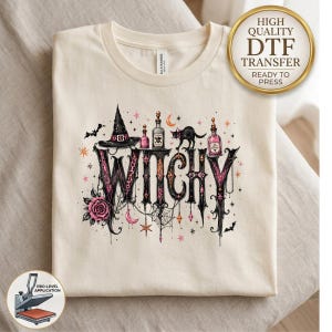 May include: A cream-colored t-shirt featuring a graphic print with the word WITCHY in large black letters. The design includes a black witch hat, a black cat, pink potion bottles, a rose, a spider web, stars, and moons in pink and black.