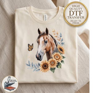 May include: A cream-colored t-shirt featuring a graphic print of a brown horse head surrounded by yellow sunflowers, blue-tinted leaves, and a monarch butterfly. The shirt includes a circular gold badge in the top right corner with the text High Quality DTF Transfer Ready To Press.