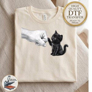 May include: A cream-colored t-shirt featuring a black and white graphic of a human hand making a fist bump gesture with a small black kitten. The design is centered on the chest area of the shirt, which has a crew neckline and short sleeves.