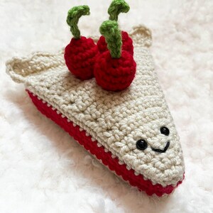 May include: A crocheted slice of pie with a red filling and two red cherries with green stems. The pie has a smiling face and is beige with a red border. The word "pattern" is at the bottom.