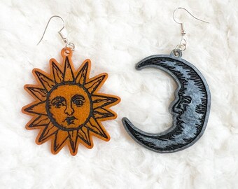 May include: A pair of celestial-themed earrings. One earring is a sun with an orange background and black details. The other is a crescent moon with a gray background and black details. Both earrings have silver-colored ear wires.
