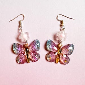 May include: A pair of butterfly earrings with iridescent wings in shades of pink, purple, and blue. Each earring features a gold-colored body and a small, clear flower with a pink center. The earrings hang from gold-colored hooks.