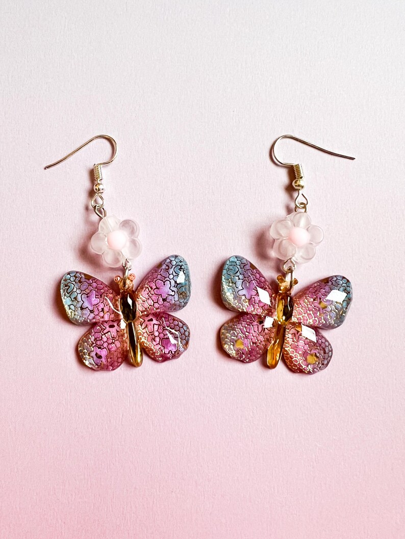 May include: A pair of butterfly earrings with iridescent wings in shades of pink, purple, and blue. Each earring features a gold-colored body and a small, clear flower with a pink center. The earrings hang from gold-colored hooks.