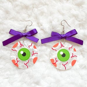 May include: A pair of Halloween earrings. Each earring has a clear, round design with a green iris, black pupil, and red veins. Orange accents are also present. A purple bow is attached to the top of each earring.