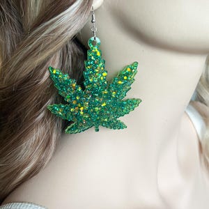 May include: A dangling earring shaped like a cannabis leaf, crafted from translucent green resin embedded with chunky gold and green glitter. The earring features a silver-toned metal hook for attachment and has a textured, sparkling finish.