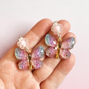 May include: A pair of butterfly earrings with iridescent wings in shades of pink, purple, and blue. Each earring features a small, light pink flower above the butterfly and gold-colored accents. The earrings are held in a hand against a pink background.