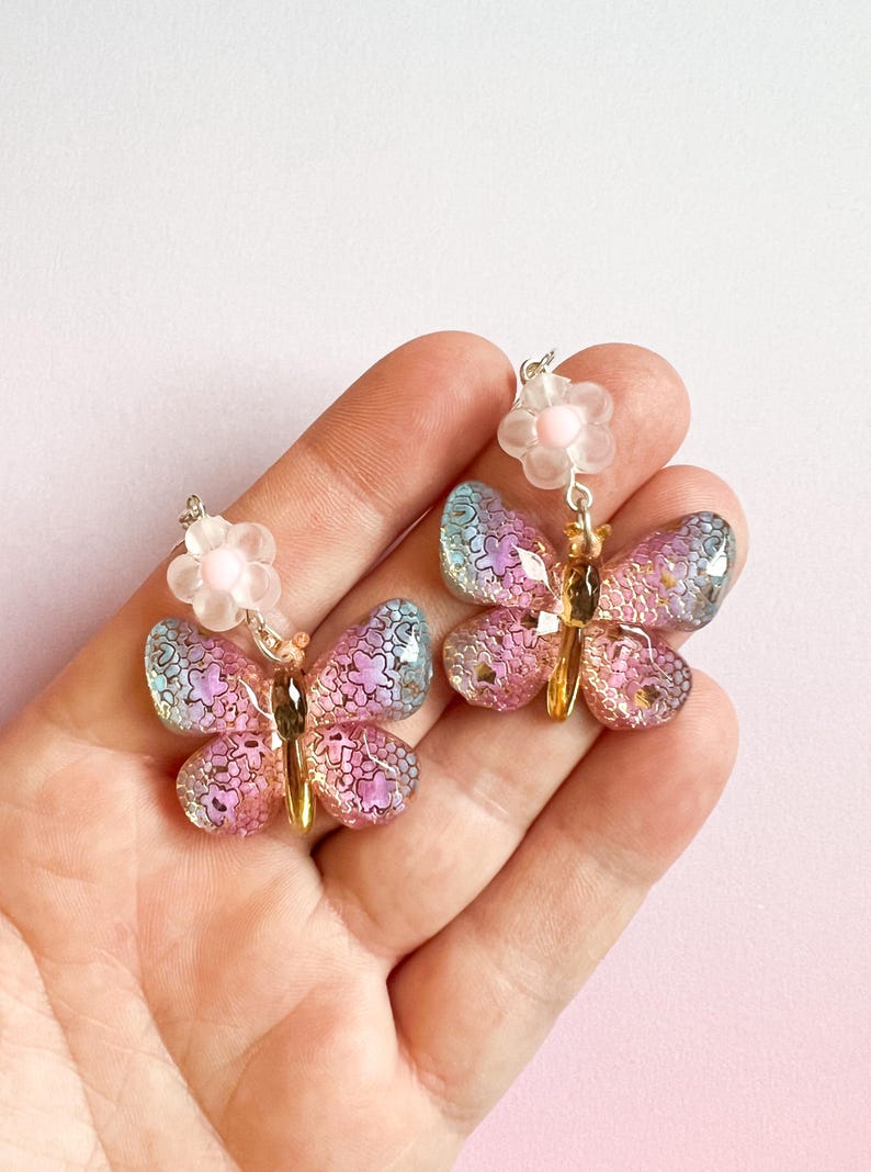 May include: A pair of butterfly earrings with iridescent wings in shades of pink, purple, and blue. Each earring features a small, light pink flower above the butterfly and gold-colored accents. The earrings are held in a hand against a pink background.