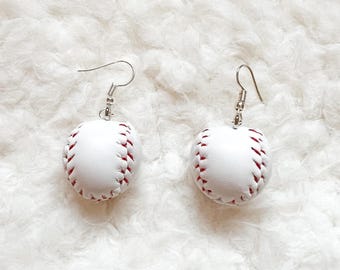 May include: A pair of baseball-themed earrings. Each earring features a white baseball with red stitching, suspended from a silver-colored fish hook. The earrings are set against a fluffy white background.