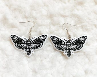 May include: A pair of white and black moth earrings. The earrings feature a detailed moth design with a skull in the center. The moth wings are black with intricate patterns. The earrings hang from silver-coloured hooks.