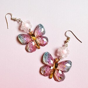 May include: A pair of butterfly earrings with iridescent wings in shades of pink, blue, and gold. Each earring features a small, white flower and a silver hook. The earrings are set against a soft pink background.
