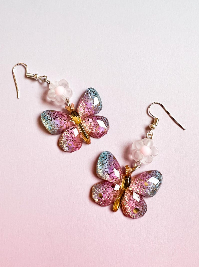 May include: A pair of butterfly earrings with iridescent wings in shades of pink, blue, and gold. Each earring features a small, white flower and a silver hook. The earrings are set against a soft pink background.