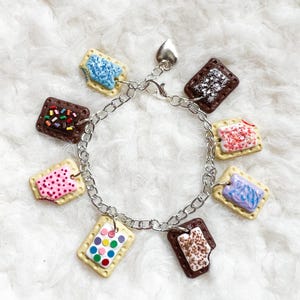 Könnte beinhalten: Ein silbernes Charm-Armband mit mehreren kleinen, bunten, rechteckigen Lebensmittel-Anhängern. Die Anhänger sind so gestaltet, dass sie glasierten Gebäckstücken mit verschiedenen Belägen wie Streuseln und Punkten ähneln. Ein herzförmiger Anhänger hängt in der Nähe des Verschlusses.